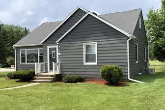 A small gray house with white trim, featuring a front porch, several windows, a gable roof, and surrounded by a well-kept lawn under a cloudy sky.