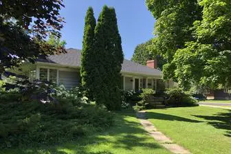 Single-story house with a gray roof and beige siding, surrounded by a lush green lawn and tall trees under a clear blue sky.