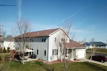 Aerial view of a two-story house with a white exterior, attached garage, and a red car parked outside, in a suburban neighborhood on a sunny day.