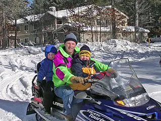 A family of three, wearing winter gear, smiles while sitting on a snowmobile in a snowy landscape with trees and a house in the background.