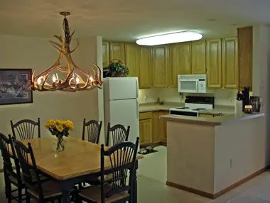 A well-lit kitchen with wooden cabinets, a dining table set, and modern appliances, featuring a hanging chandelier and a floral centerpiece.