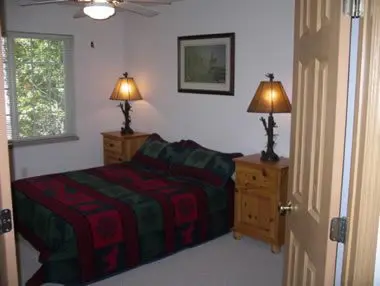 A small, cozy bedroom with a single bed covered in a red and green quilt, wooden nightstands with lamps, and a framed picture above the bed.