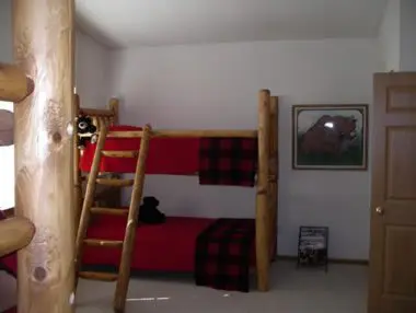 A rustic bedroom featuring a log-framed bunk bed with red plaid bedding, a framed bear painting on the wall, and a wooden door on the right.