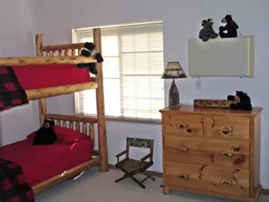 A bedroom featuring a wooden bunk bed with red bedding, a dresser, and stuffed animals arranged on the furniture and window sill.