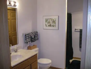 A small bathroom with a sink, mirror, wooden cabinet, toilet, and a partially visible shower with hanging towels. a framed floral picture hangs on the wall.