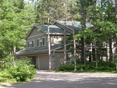 Two-story house with a gray exterior and green roof, nestled among tall pine trees, featuring a double garage and a paved driveway.