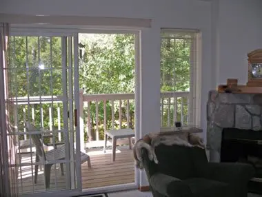 Living room with a green chair and a fireplace, overlooking a deck with chairs through large sliding glass doors and windows, surrounded by trees.