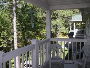 A cozy porch with white railings, furnished with chairs, overlooking dense green woods.