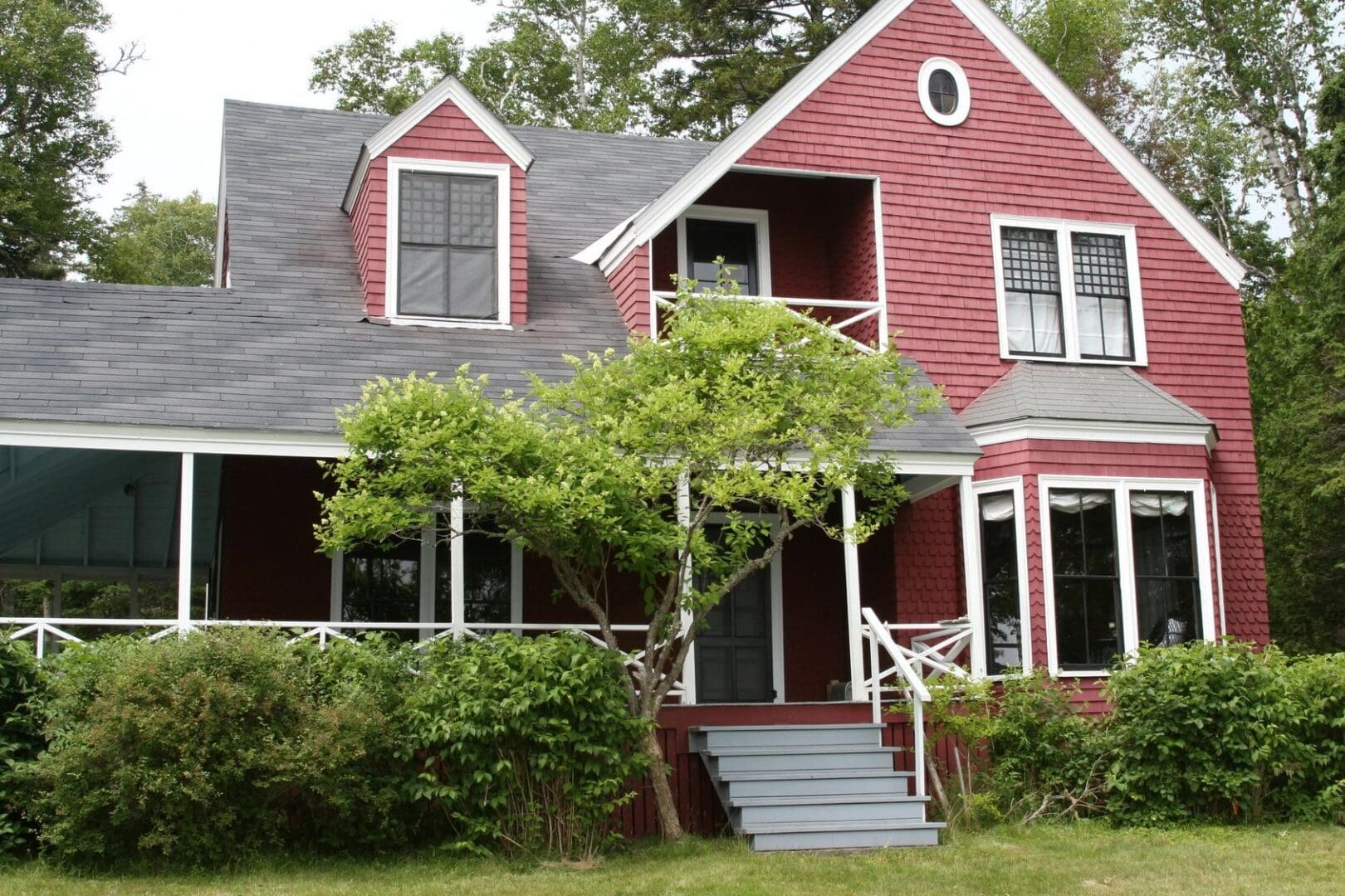 Red two-story house with white trim, a covered porch, and a surrounding green lawn with a small tree in front.