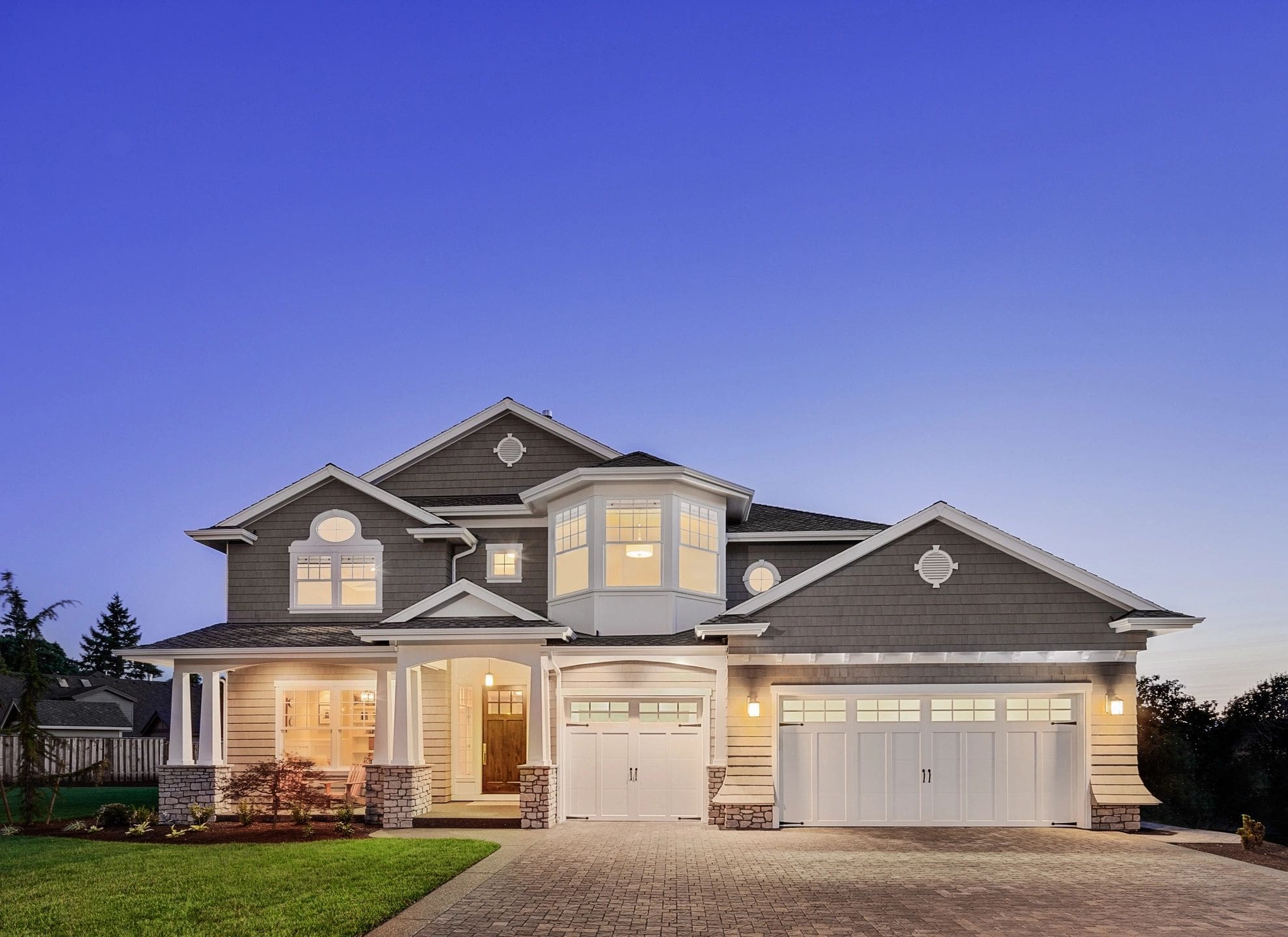 Large two-story house with three-car garage and illuminated windows, set against a twilight sky.