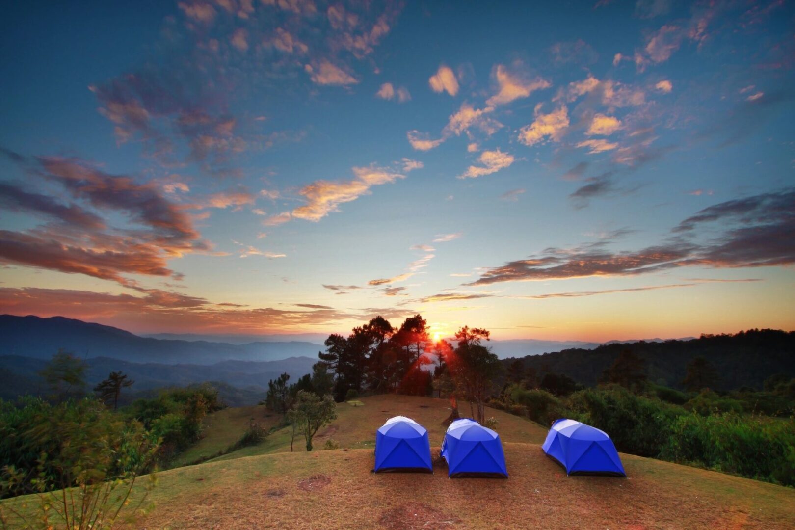 Three blue tents pitched on a hill overlooking a scenic sunset with mountains in the background and a colorful sky.