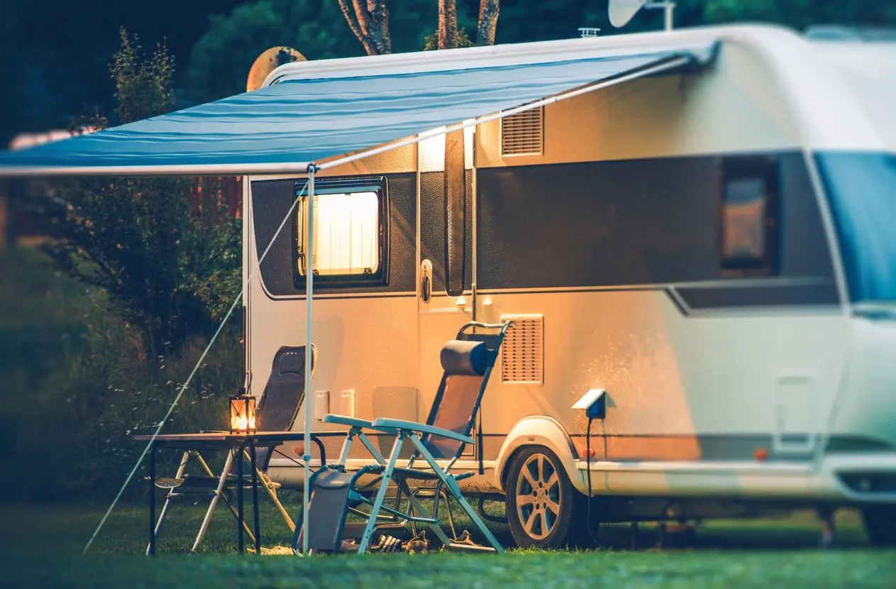 A peaceful evening at a campsite with a caravan, an open awning, a lit lantern on a table, and camping chairs set up on grass.