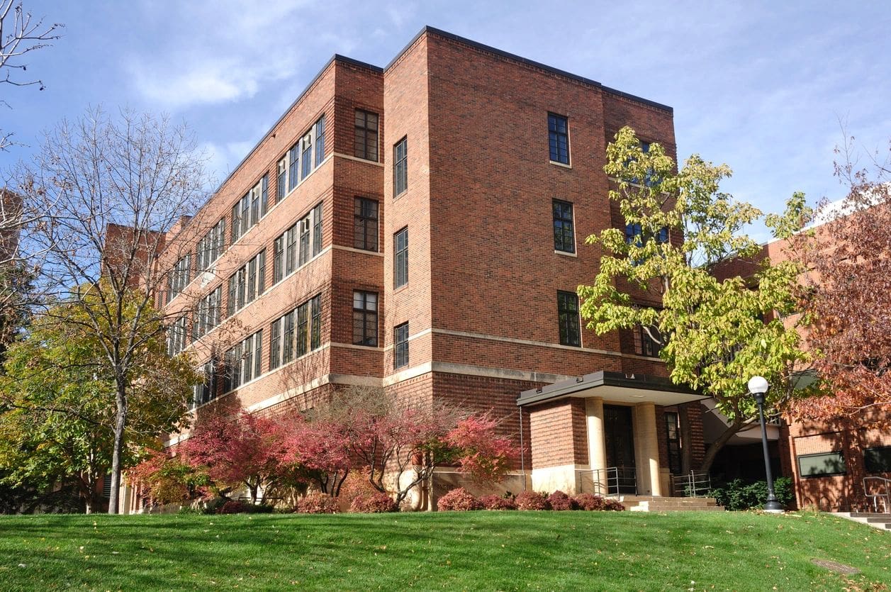 A modern brick university building surrounded by green lawns and autumn-colored trees under a clear sky.