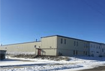 A two-story industrial building with a gray exterior, under a clear blue sky, surrounded by patches of snow.