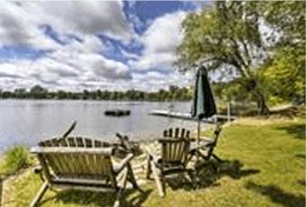 Two wooden chairs face a tranquil lake with a small boat floating, under a clear sky and next to a closed umbrella.