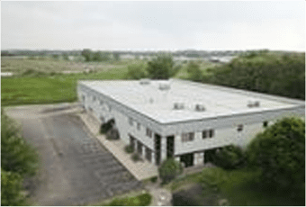 Aerial view of a two-story rectangular building with a flat roof, surrounded by green fields and a partly visible parking area.