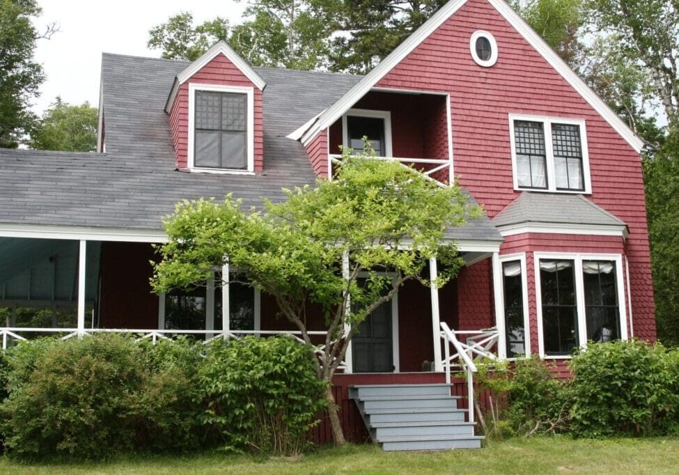 Red two-story house with white trim, a covered porch, and a surrounding green lawn with a small tree in front.