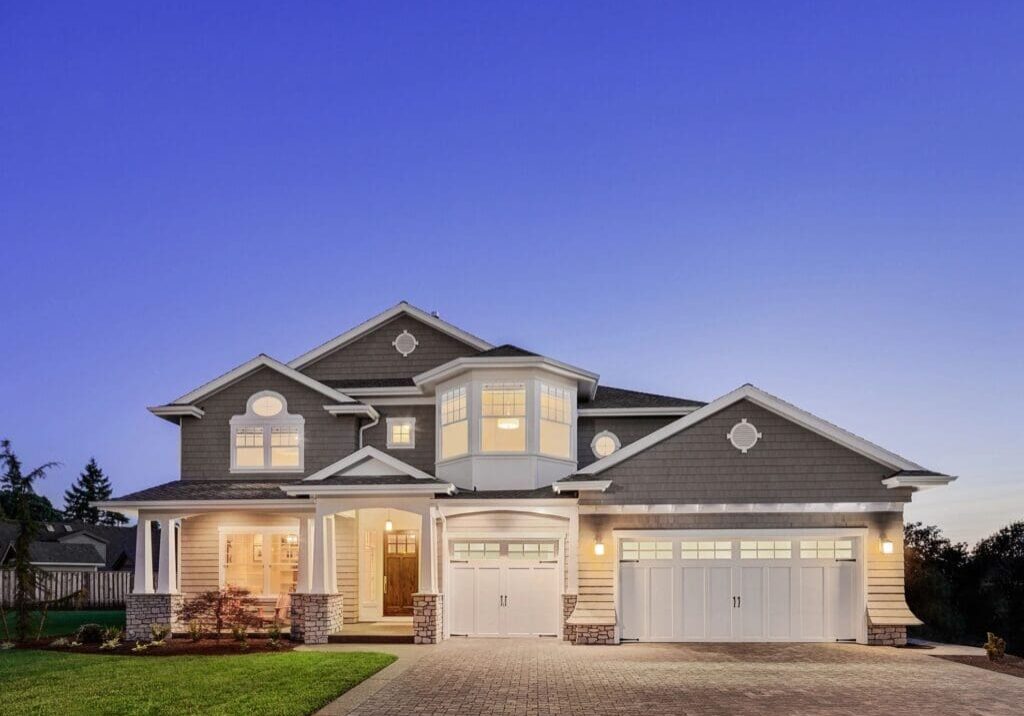 Large two-story house with three-car garage and illuminated windows, set against a twilight sky.