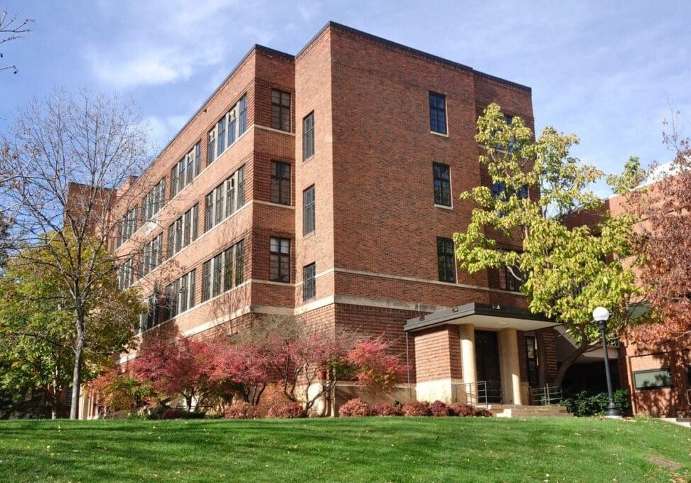 A modern brick university building surrounded by green lawns and autumn-colored trees under a clear sky.